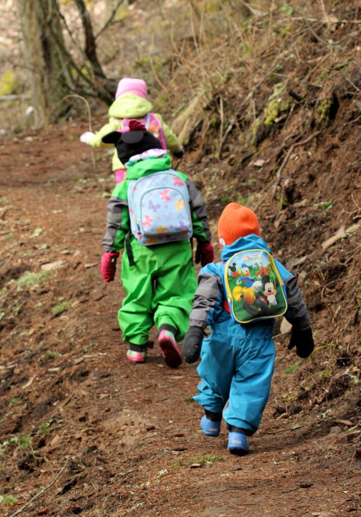 Three young kids hiking up a dirt trail wearing backpacks and rainsuits.