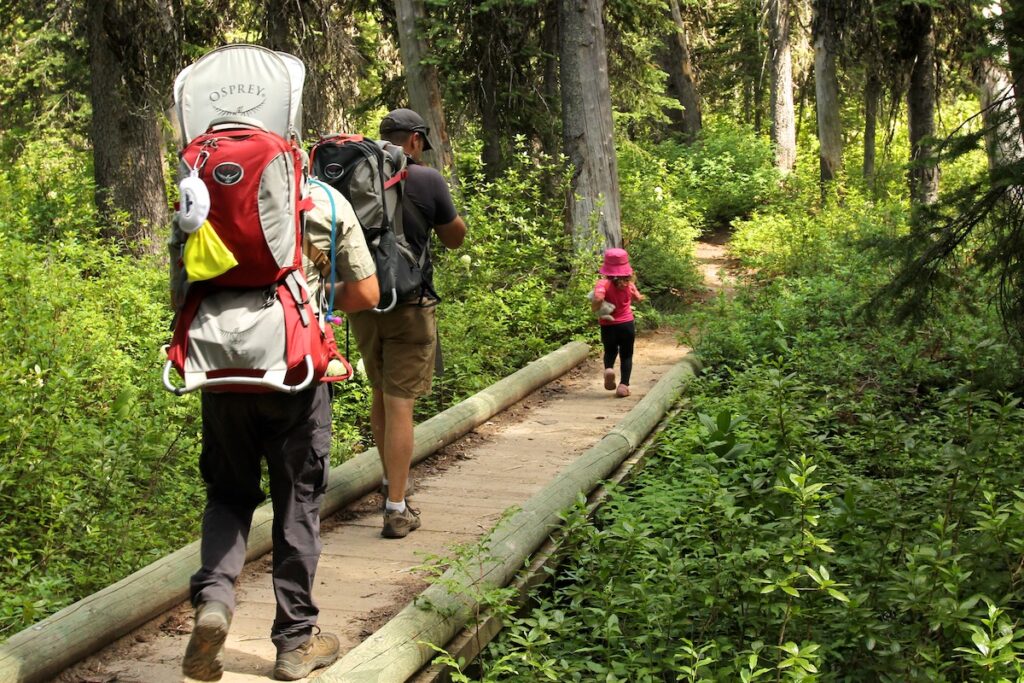 Two dads walking across a boardwalk in the woods carrying child carrier backpacks. Toddler girl in front of them leading the way.